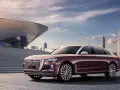 Elegancki, bordowy Hongqi H9 z chromowanymi detalami stoi przed nowoczesnym budynkiem o falistej fasadzie, w tle miejski krajobraz.