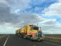 Żółta ciężarówka typu road train z naczepami na pustej drodze pod zachmurzonym niebem.