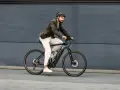 Mężczyzna w kasku i marynarce jedzie na rowerze elektrycznym po szarym tle.
