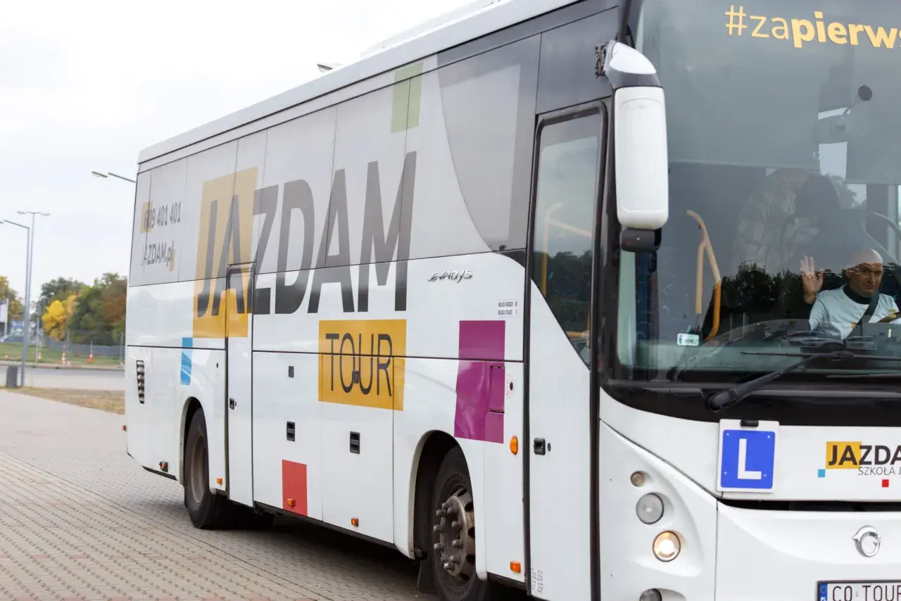 Biały autokar z napisem "JAZDAM TOUR" i hasłem "#zapierw". Kierowca macha. Zastanawiasz się, ile kosztuje autobus?
