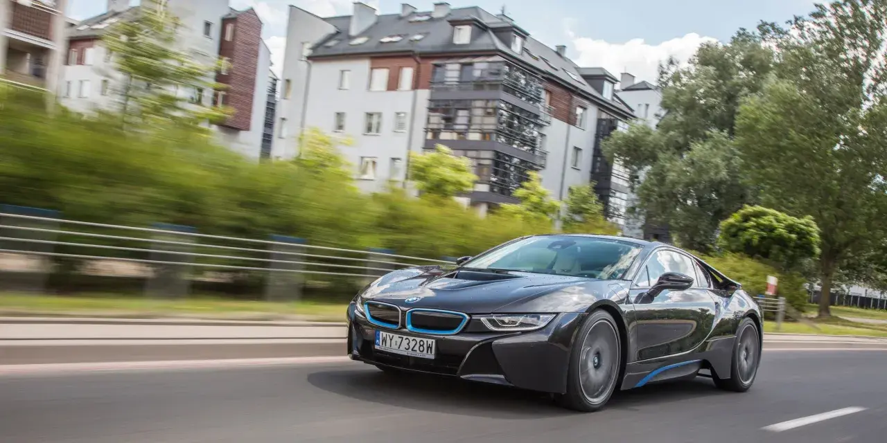 Czarny BMW i8 pędzi ulicą, mijając nowoczesne budynki i zielone drzewa.