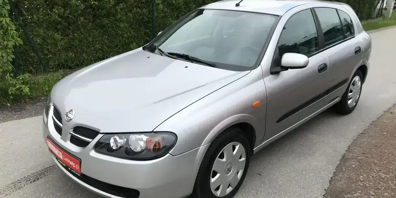 Srebrny Nissan Almera hatchback stoi na asfaltowej drodze, otoczony zielenią.