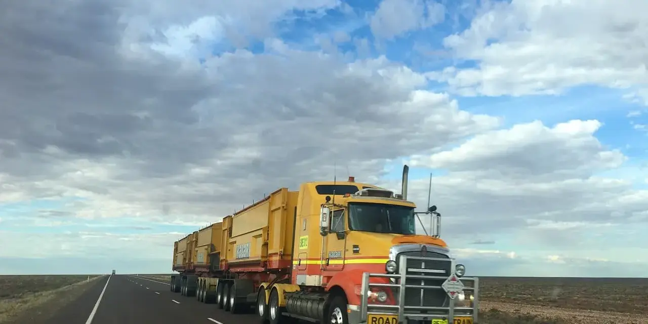 Żółta ciężarówka typu road train z naczepami na pustej drodze pod zachmurzonym niebem.