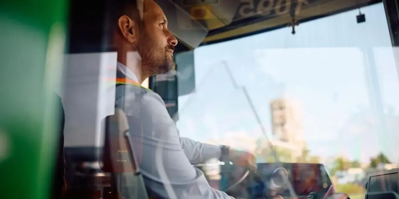 Skupiony kierowca autobusu prowadzi pojazd, trzymając kierownicę. Widać jego profil i fragment deski rozdzielczej.