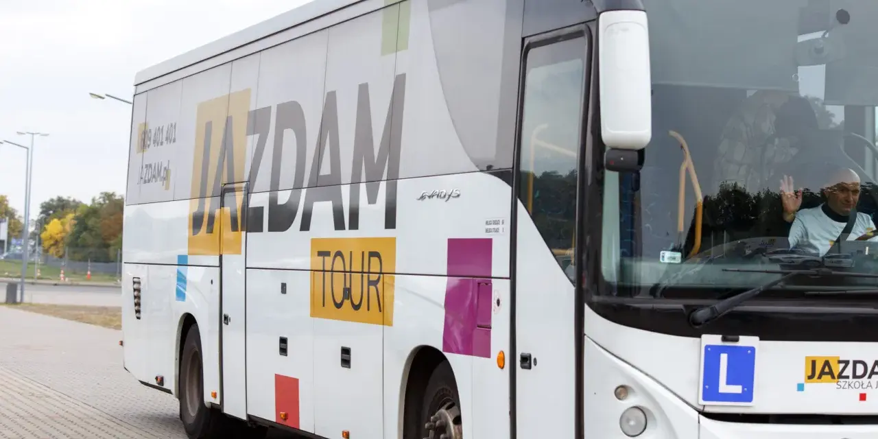 Biały autokar z napisem "JAZDAM TOUR" i hasłem "#zapierw". Kierowca macha. Zastanawiasz się, ile kosztuje autobus?