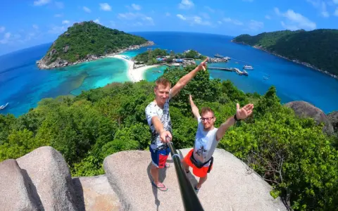Dwóch mężczyzn robi selfie na tle tropikalnej wyspy z turkusową wodą i plażą.