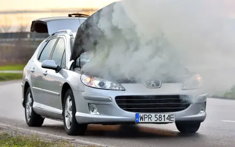 Srebrny samochód Peugeot z otwartą maską, z której wydobywa się gęsty dym.