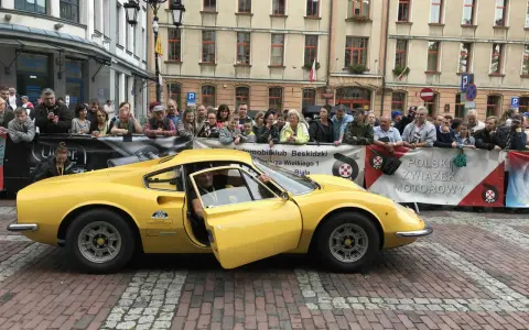 Żółte Ferrari Dino z otwartymi drzwiami, kierowca w środku, tłum widzów za barierką.