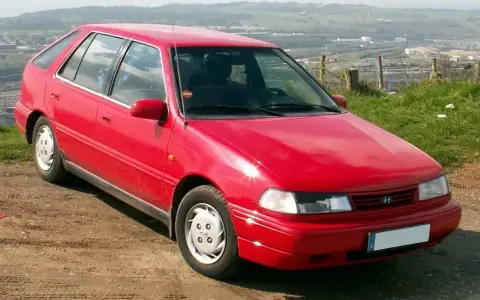 Czerwony Hyundai Pony z lat 90. stoi na polnej drodze, z widokiem na dolinę i miasto w tle.