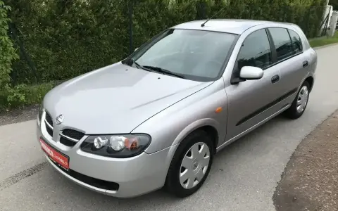 Srebrny Nissan Almera hatchback stoi na asfaltowej drodze, otoczony zielenią.