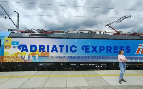 Kobieta stoi obok lokomotywy Adriatic Express z widokiem na morze i żaglówki.