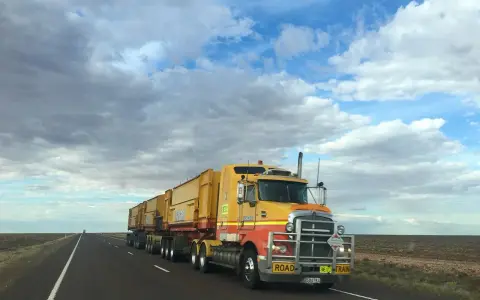 Żółta ciężarówka typu road train z naczepami na pustej drodze pod zachmurzonym niebem.