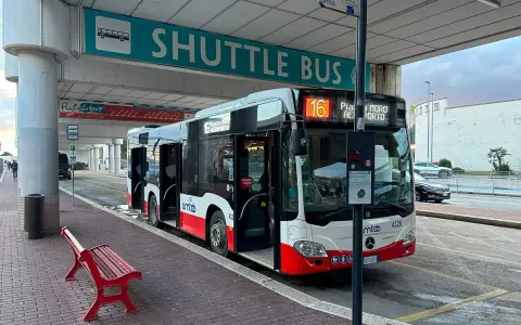 Autobus wahadłowy z otwartymi drzwiami na przystanku lotniskowym, z napisem "SHUTTLE BUS".