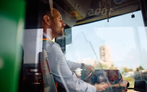 Skupiony kierowca autobusu prowadzi pojazd, trzymając kierownicę. Widać jego profil i fragment deski rozdzielczej.