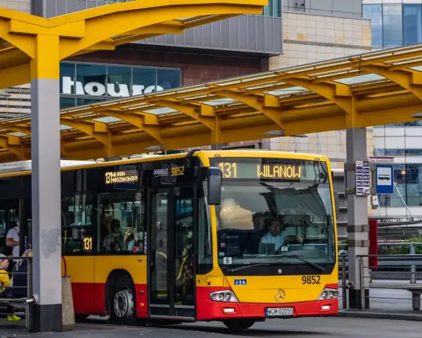 Żółto-czerwony autobus miejski na przystanku. Kierowca autobusu, jak i pasażerowie, zastanawiają się, ile zarabia kierowca autobusu.