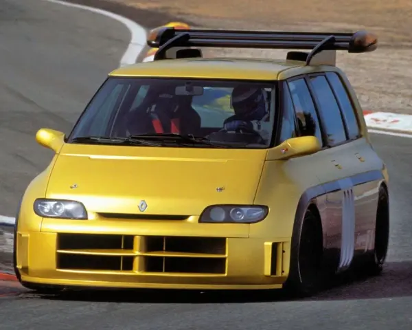 Żółty Renault Espace F1 z kierowcą w kasku na torze wyścigowym. Potężny spojler dachowy i agresywny wygląd.
