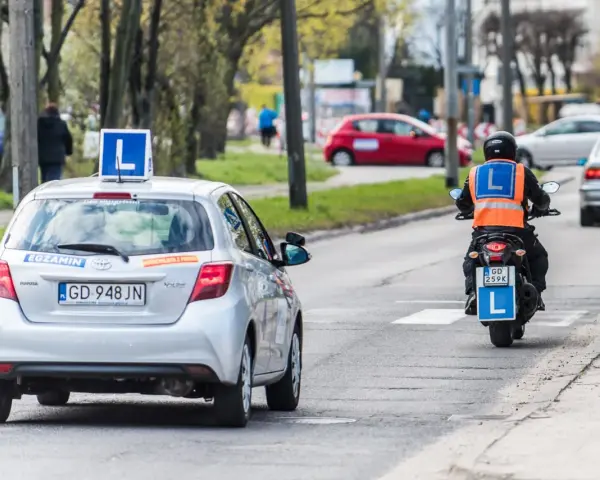 Srebrny samochód z literą "L" na dachu i motocyklista w kamizelce odblaskowej, obaj z literą "L" z tyłu, poruszają się po drodze. To kategoria a nauki jazdy.