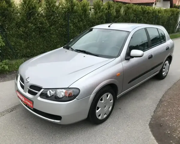 Srebrny Nissan Almera hatchback stoi na asfaltowej drodze, otoczony zielenią.
