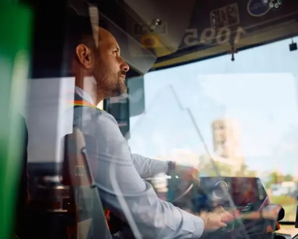 Skupiony kierowca autobusu prowadzi pojazd, trzymając kierownicę. Widać jego profil i fragment deski rozdzielczej.