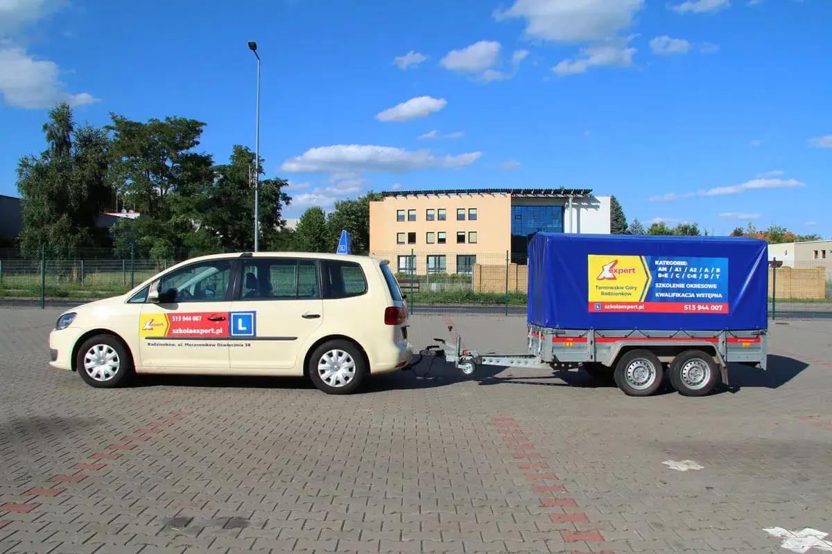 Jasny samochód z przyczepą szkoleniową do nauki jazdy kat. B+E. Na przyczepie logo szkoły jazdy i kategorie uprawnień.