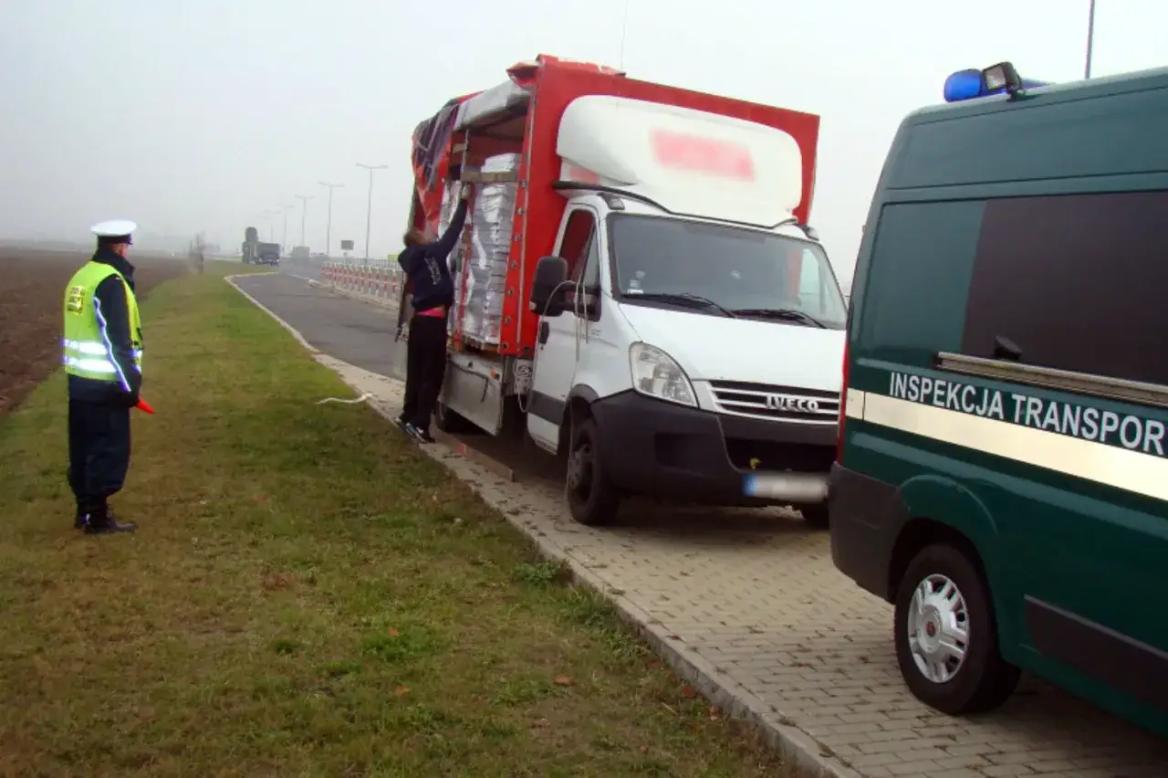 Inspektor drogowy kieruje ruchem przy białej ciężarówce, podczas gdy pracownik sprawdza ładunek. Obok stoi zielony furgon z napisem 