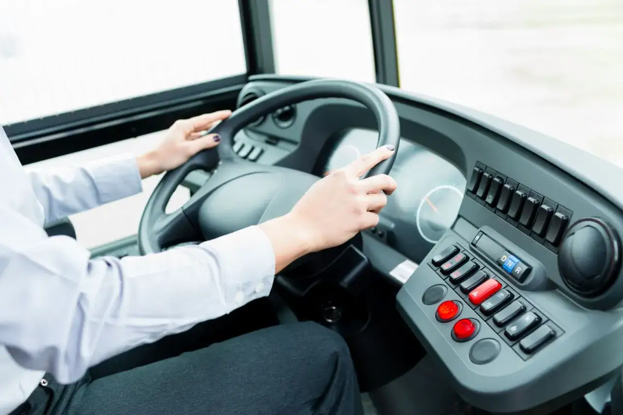 Ręce kierowcy autobusu na kierownicy. Widoczny panel sterowania z przyciskami i wyświetlaczem.