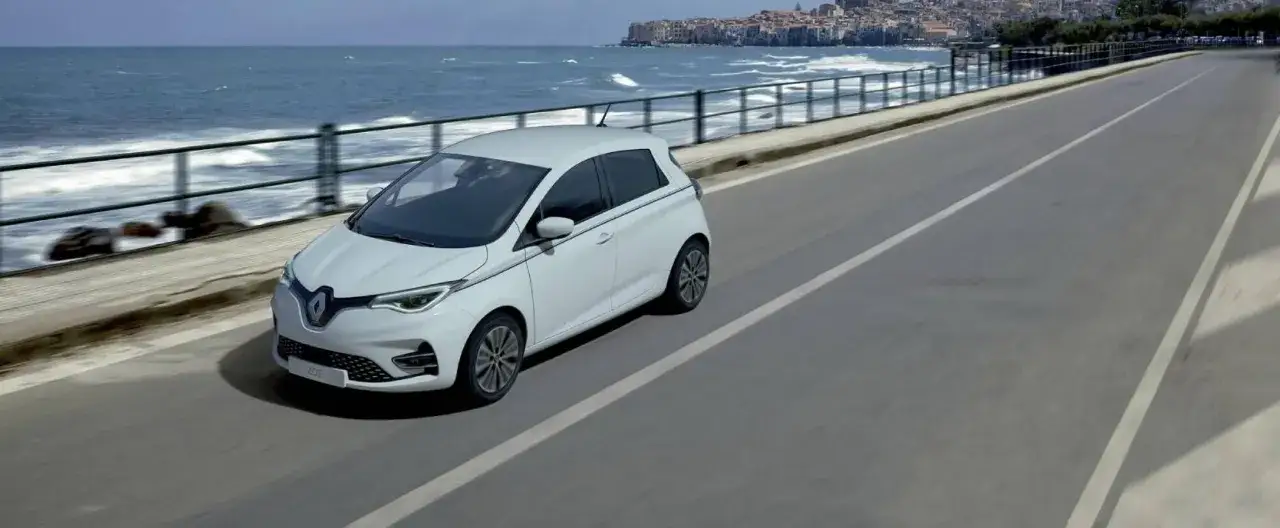 Biały Renault Zoe jedzie wzdłuż wybrzeża z widokiem na miasto i morze.