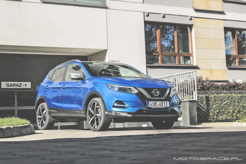 Błękitny Nissan Qashqai 2018 zaparkowany przed nowoczesnym budynkiem z garażem.