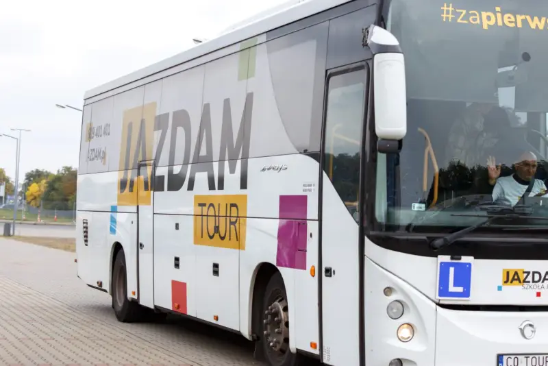 Biały autokar z napisem "JAZDAM TOUR" i hasłem "#zapierw". Kierowca macha. Zastanawiasz się, ile kosztuje autobus?